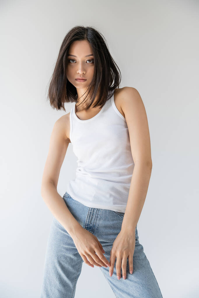 brünette asiatische Frau in weißem Tank-Top und blauen Jeans schaut in die Kamera isoliert auf grau - Foto, Bild