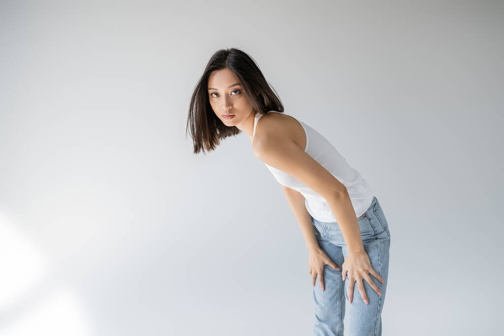 junge und schlanke asiatische Frau in Jeans und Tank-Top schaut in die Kamera auf grauem Hintergrund - Foto, Bild