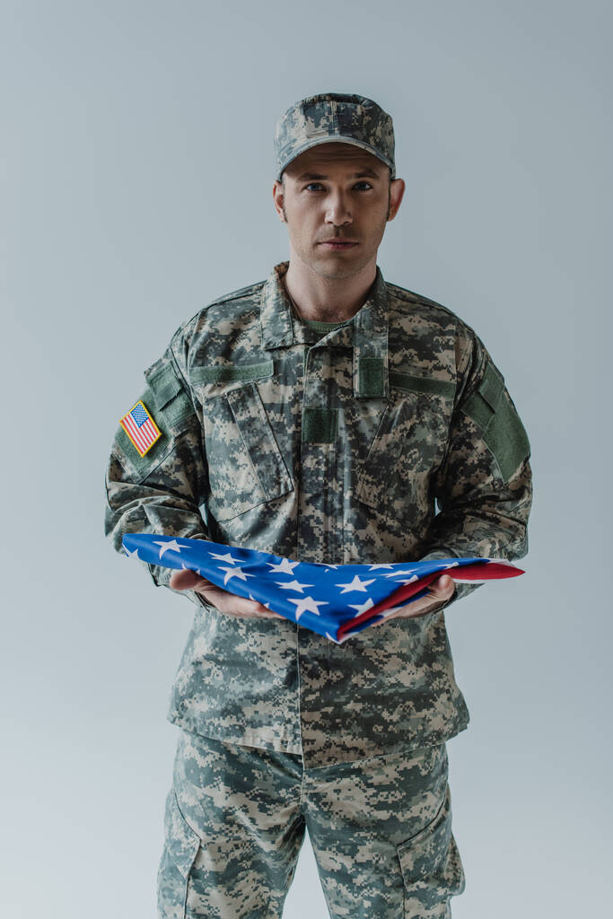 Soldat américain en uniforme tenant le drapeau plié des États-Unis pendant la journée commémorative isolé sur gris  - Photo, image