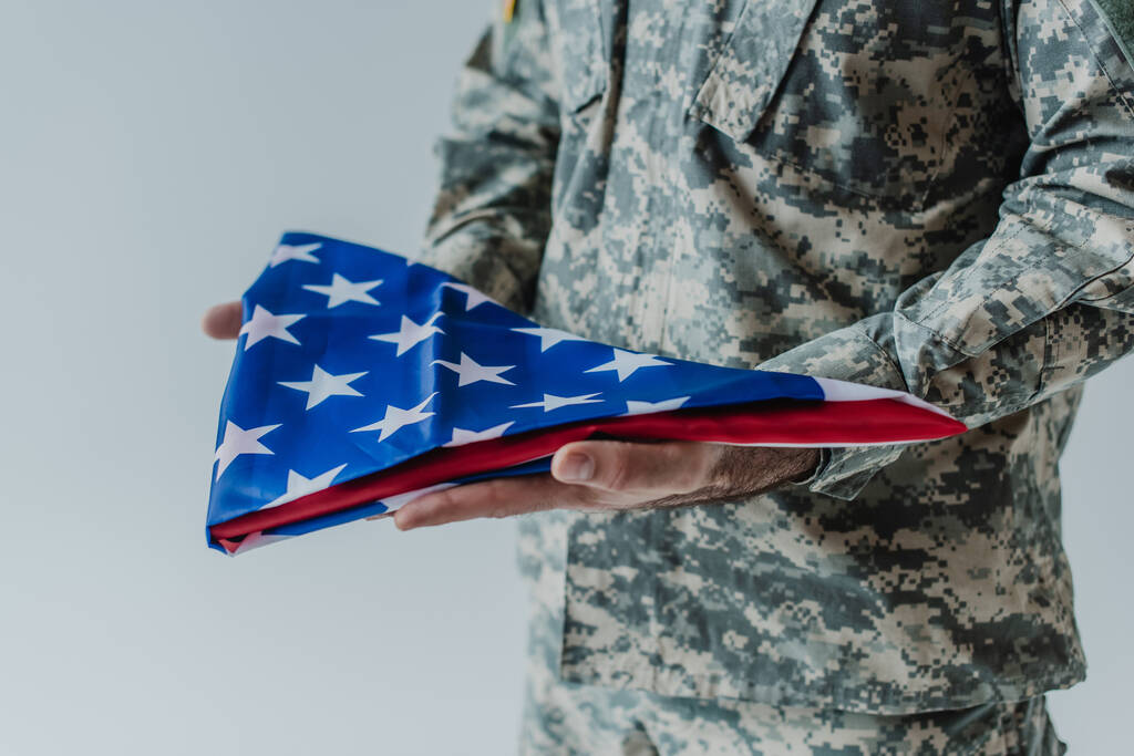 vista parziale del soldato americano in uniforme con bandiera piegata degli Stati Uniti durante il giorno commemorativo isolato sul grigio  - Foto, immagini