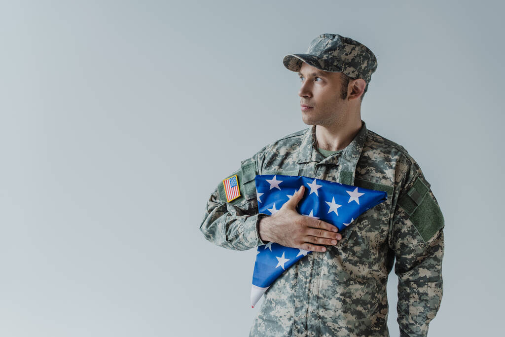 soldado americano de boa aparência em uniforme militar segurando bandeira dobrada dos Estados Unidos durante o dia comemorativo isolado em cinza  - Foto, Imagem