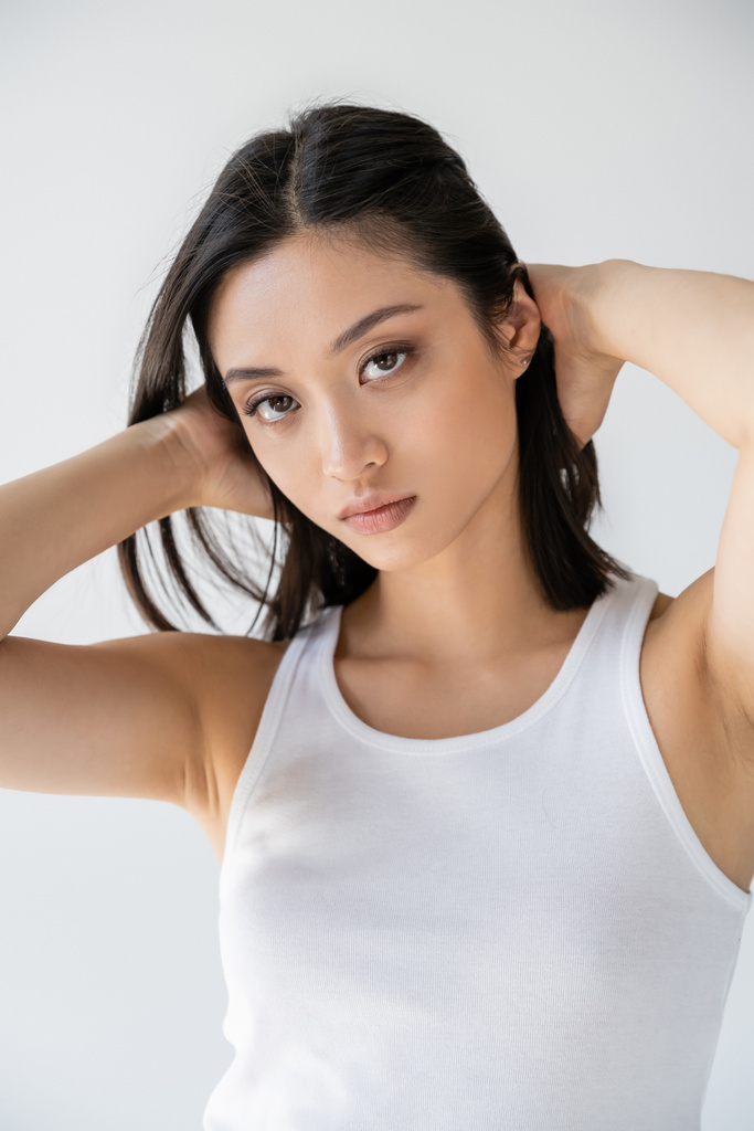 jeune asiatique femme en blanc débardeur toucher brunette cheveux et regarder caméra isolé sur gris - Photo, image