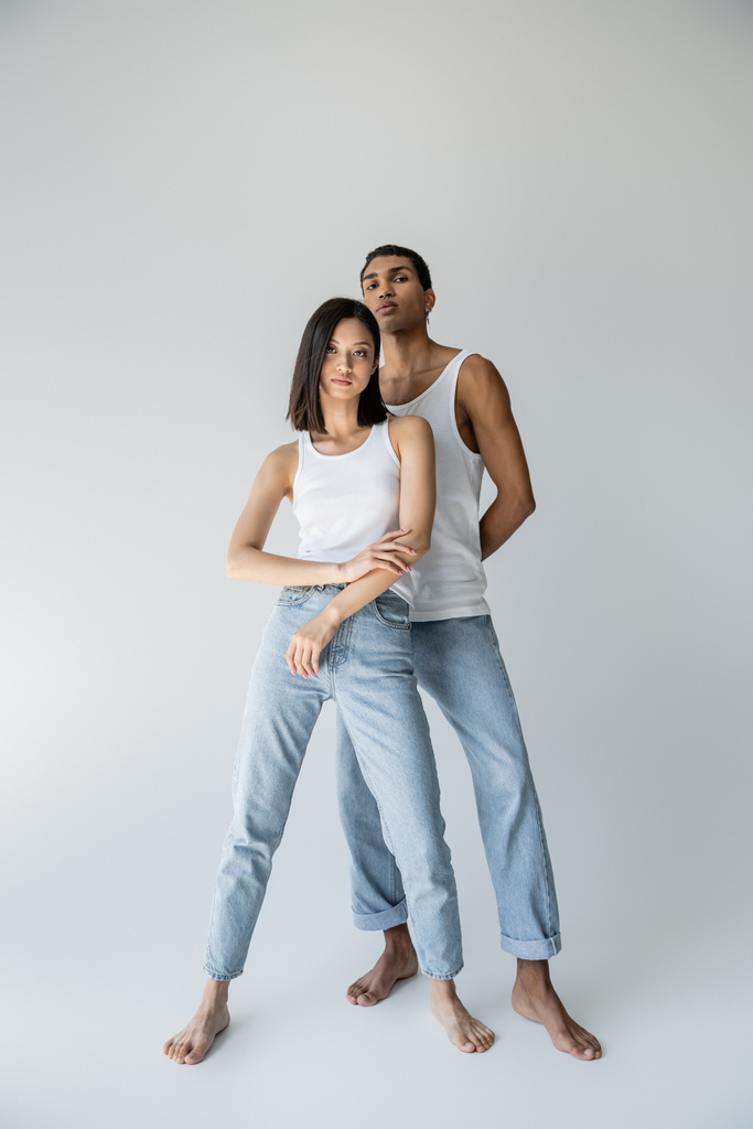 full length of barefoot and slender multiethnic couple in tank tops and jeans on grey background - Photo, Image