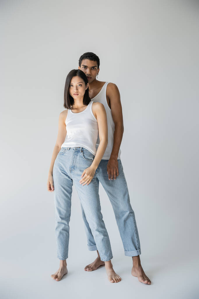 piena lunghezza di giovane e scalza coppia interrazziale in posa in canottiere bianche e jeans blu su sfondo grigio - Foto, immagini