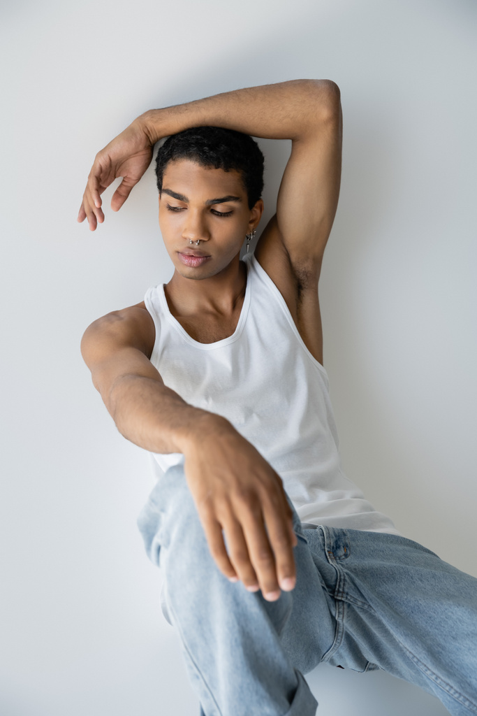 young african american guy in white tank top and jeans sitting on grey background - Photo, Image