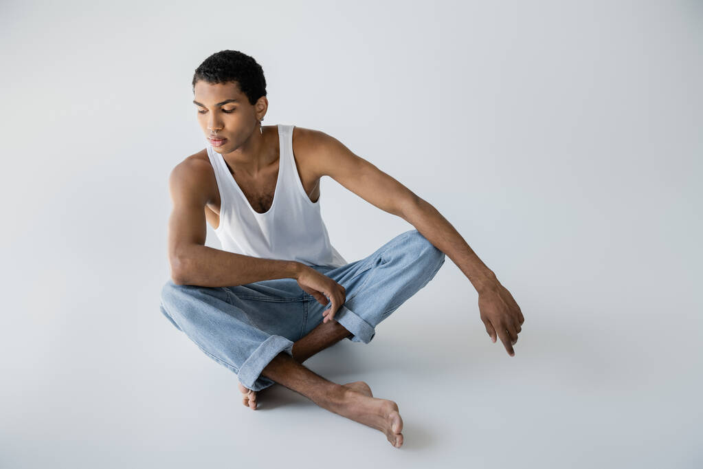 descalço afro-americano homem em jeans sentado com pernas cruzadas no fundo cinza - Foto, Imagem