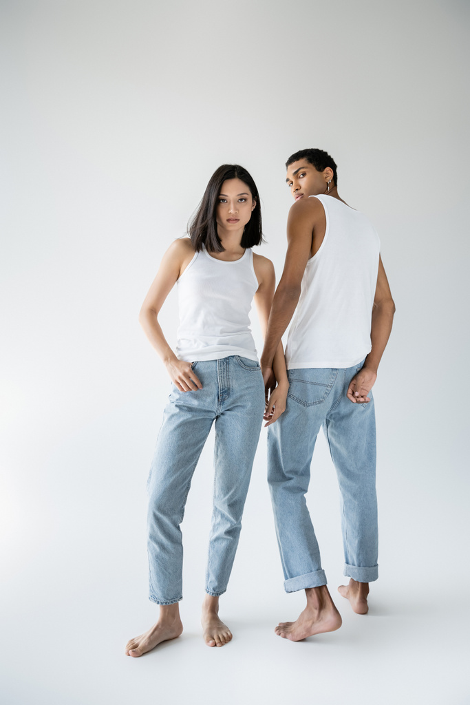 comprimento total de descalço asiático mulher posando com polegar no bolso de azul jeans perto africano americano cara no cinza fundo - Foto, Imagem