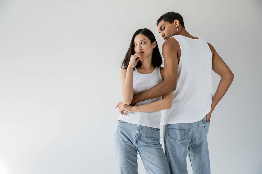 african american man in white tank top and jeans embracing dreamy asian woman looking away isolated on grey - Foto, Imagem