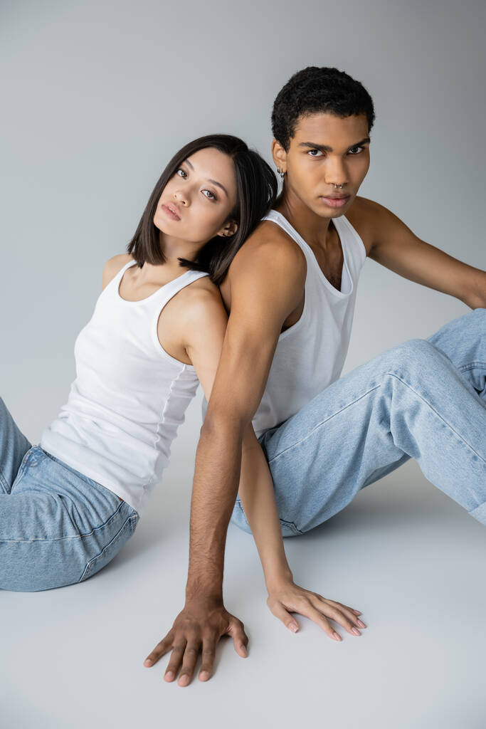 stylisches multiethnisches Paar in Blue Jeans und Tank Tops, Rücken an Rücken auf grauem Hintergrund sitzend - Foto, Bild