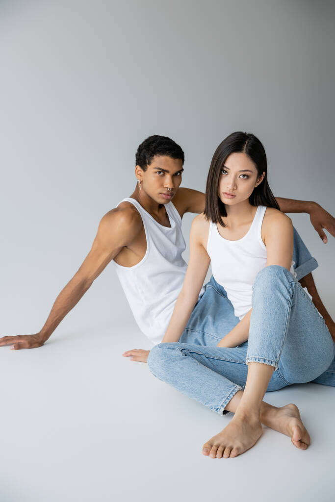 Trendy interracial Paar in Jeans und Tank-Tops sitzt und schaut in die Kamera auf grauem Hintergrund - Foto, Bild
