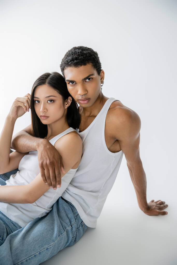 Beyaz kolsuz tişörtlü Afro-Amerikan erkeği Asyalı kadını kucaklıyor ve gri arka planda kameraya bakıyor. - Fotoğraf, Görsel