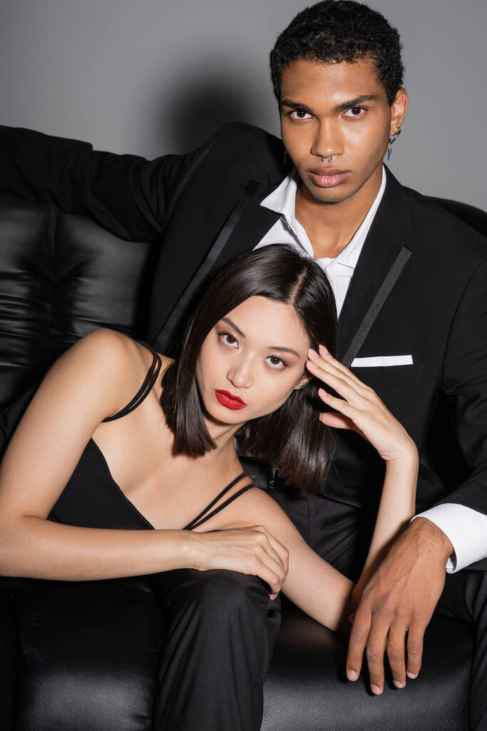 молодая и стильная межрасовая пара позирует на черном кожаном диване и смотрит в камеру на сером фоне - Фото, изображение