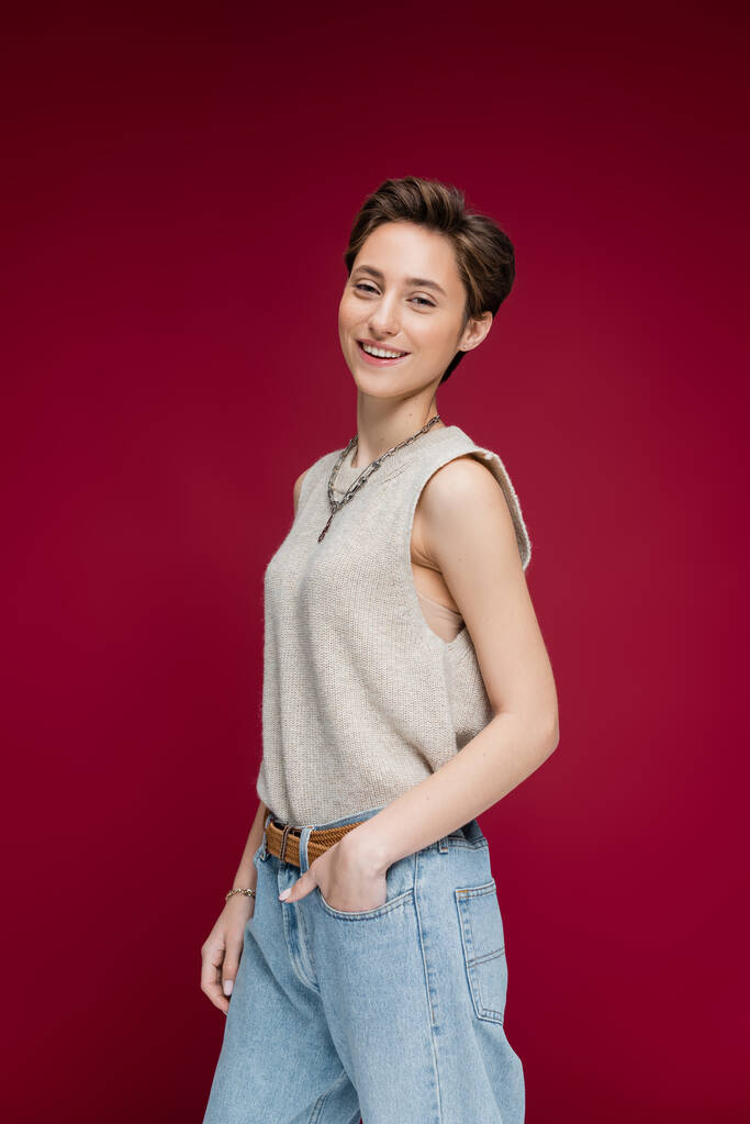 charming young woman with short hair posing with hand in pocket on dark red background - Photo, Image