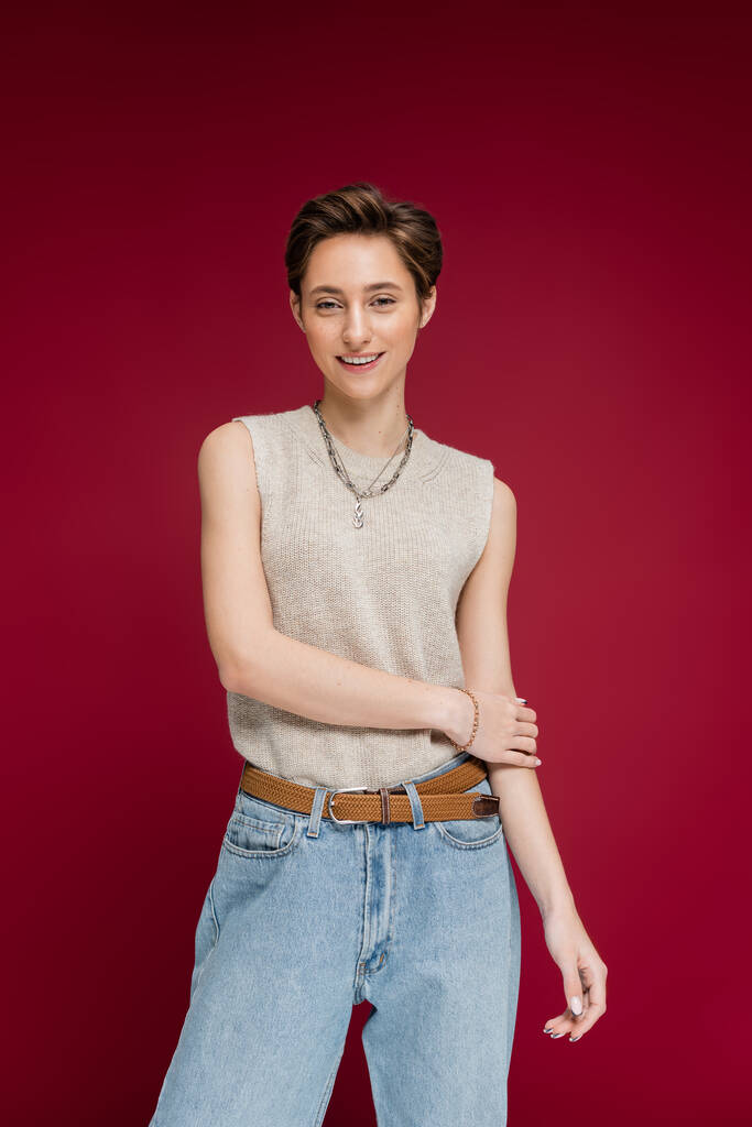 overjoyed young woman with short hair posing in jeans and sleeveless shirt on dark red background - Photo, Image