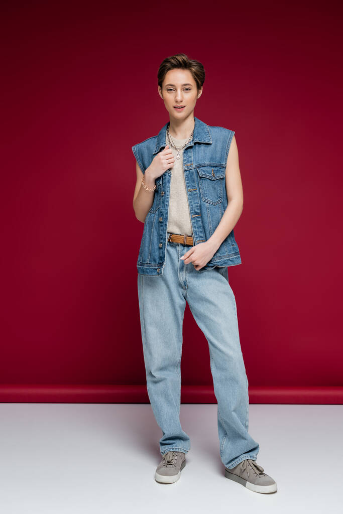 full length of stylish young woman with short hair posing in denim outfit on dark red background - Photo, Image