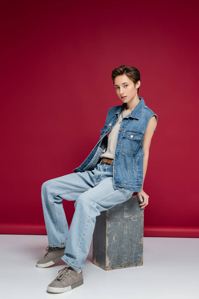 full length of pretty young model with short hair sitting in denim outfit on dark red background - Photo, Image