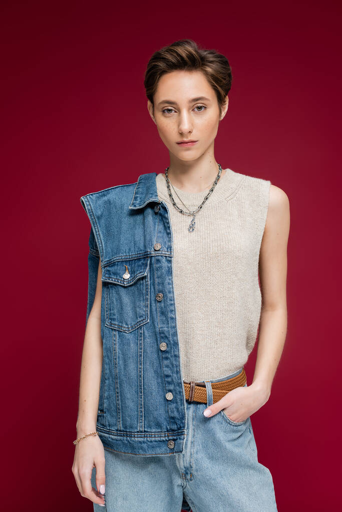 hübsches junges Model in Jeansweste stehend mit Hand in Tasche auf dunkelrotem Hintergrund - Foto, Bild