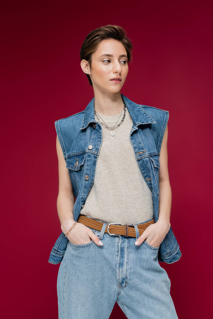 young model in denim vest standing with hands in pockets on dark red background - Photo, Image