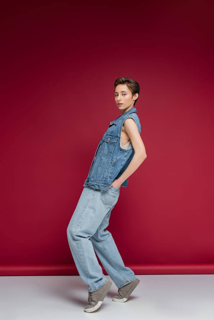 volledige lengte van stijlvol model in denim outfit met vest poserend met handen in achterzakken op jeans op bordeauxrode achtergrond  - Foto, afbeelding