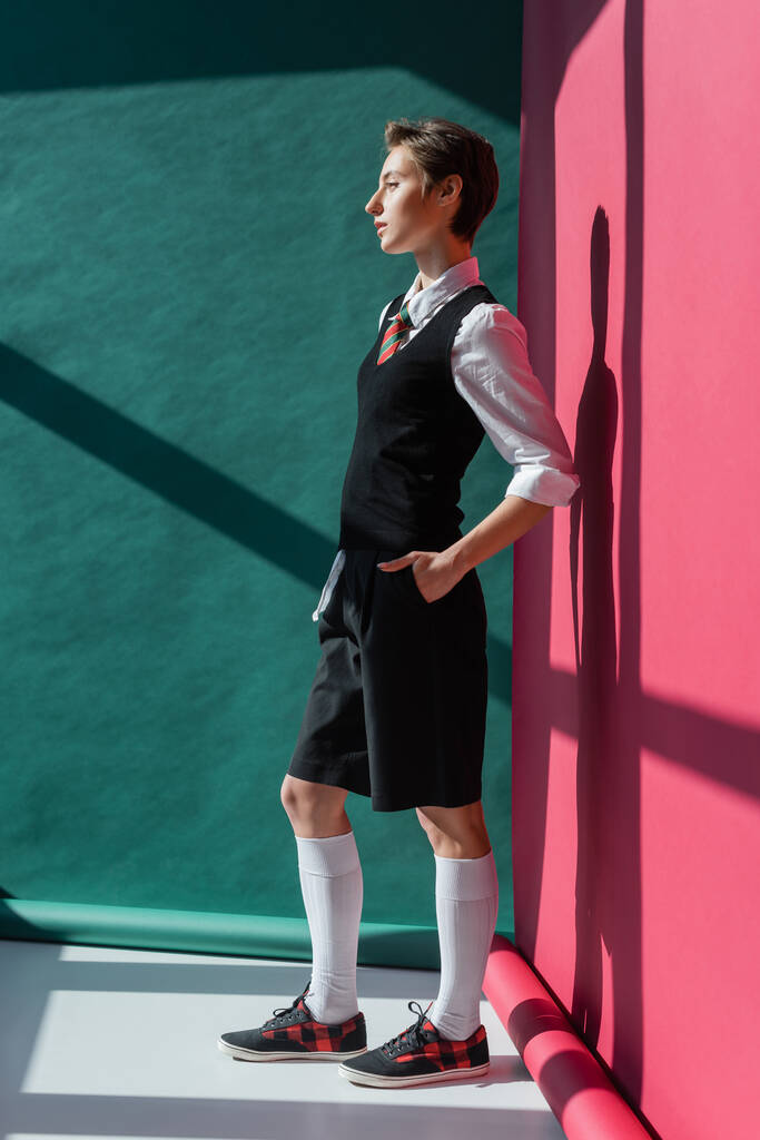 longitud completa de la elegante joven estudiante con el pelo corto posando con la mano en el bolsillo de pantalones cortos negros en rosa y verde  - Foto, imagen