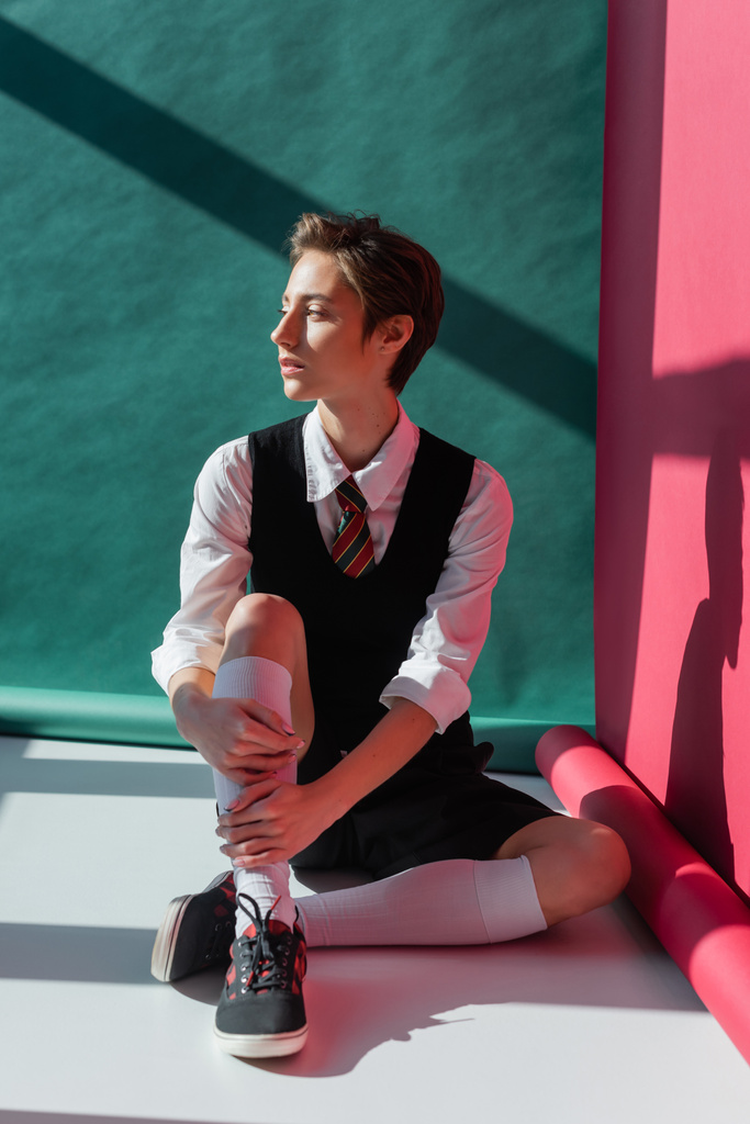 full length of stylish young student with short hair sitting in school uniform on pink and green  - Photo, Image