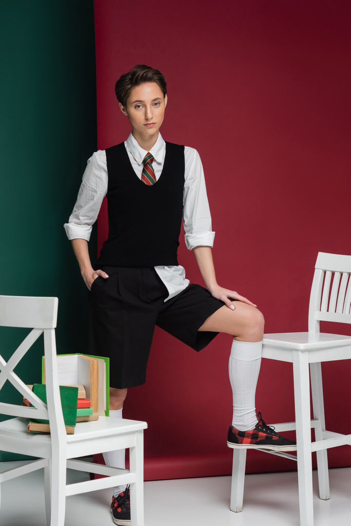stylowa młoda kobieta z krótkimi włosami pozowanie w mundurze szkolnym wokół krzeseł i książek na zielonym i różowym tle  - Zdjęcie, obraz