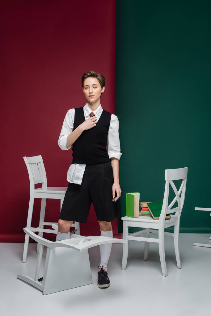повна довжина стильного студента з коротким волоссям, що тримає книгу і стоїть біля стільців на зеленому та червоному тлі
  - Фото, зображення