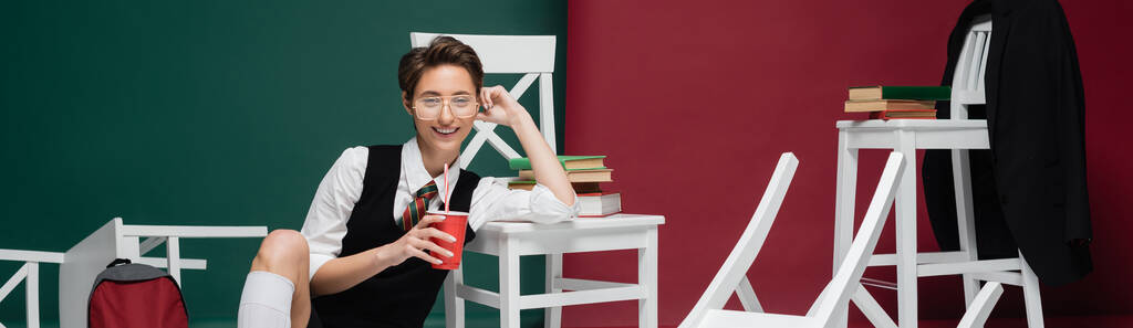 fröhliche junge Studentin in Brille mit Getränk in Plastikbecher in der Nähe von Stühlen und Büchern auf grünem und weinrotem Hintergrund, Banner  - Foto, Bild