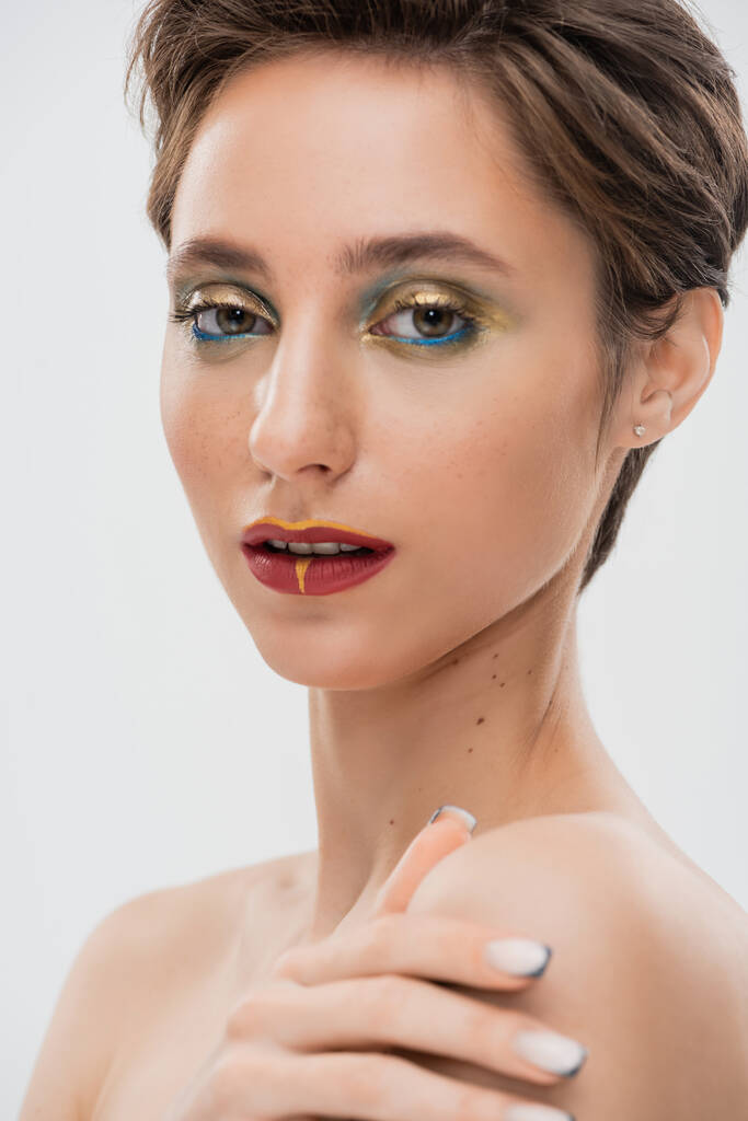 Porträt eines jungen Models mit hellem Make-up, das die nackte Schulter berührt und isoliert auf grau in die Kamera blickt  - Foto, Bild