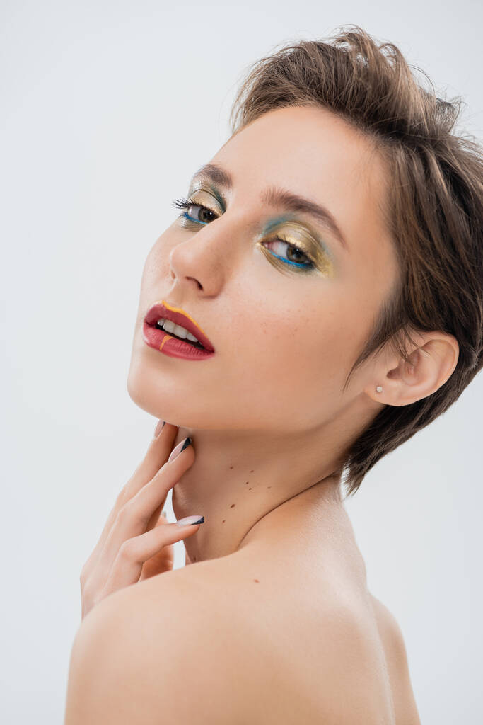 brunette young woman with shimmery eye makeup and short hair touching neck isolated on grey  - Photo, Image