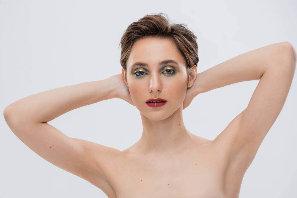 junge Frau mit schimmerndem Augen-Make-up, die Hände hinter dem Hals isoliert auf grau  - Foto, Bild