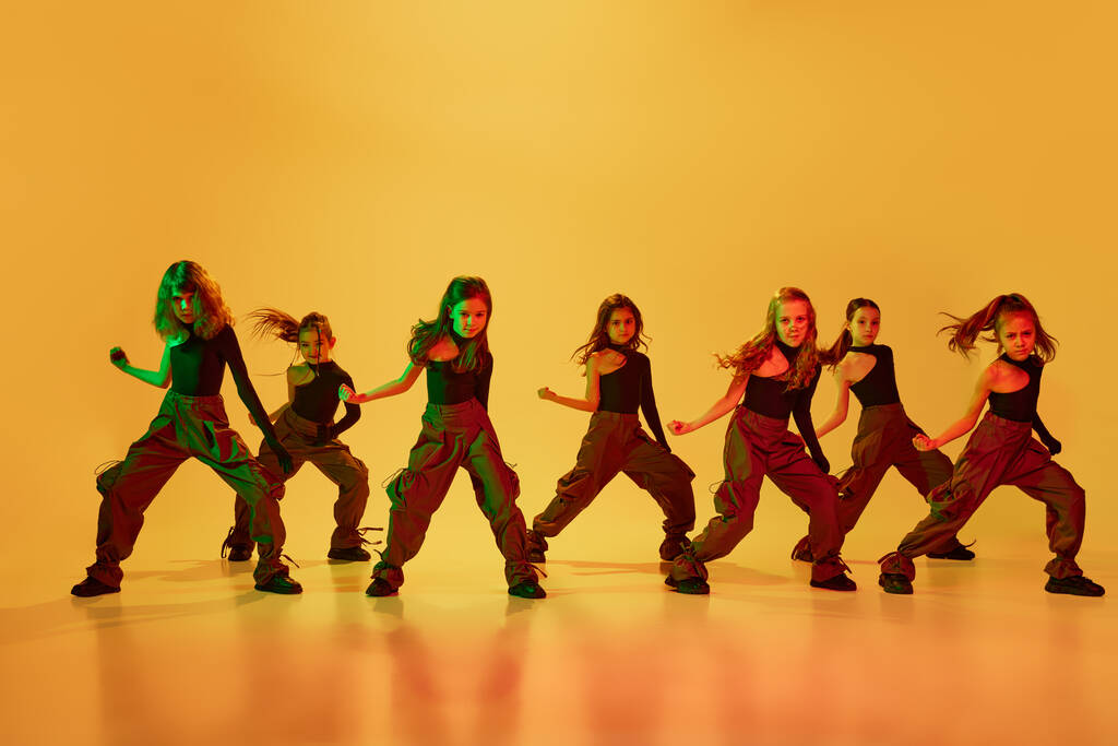 小さな才能のある女の子のグループ、ネオンの光の中で黄色のスタジオの背景にヒップホップを踊るスポーツスタイルの服の子供たち。協調運動だ。子供時代、趣味、スポーツライフスタイルの概念 - 写真・画像