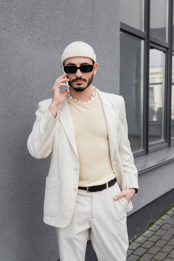 Модный гей в солнечных очках и бежевом костюме разговаривает по смартфону на улице - Фото, изображение