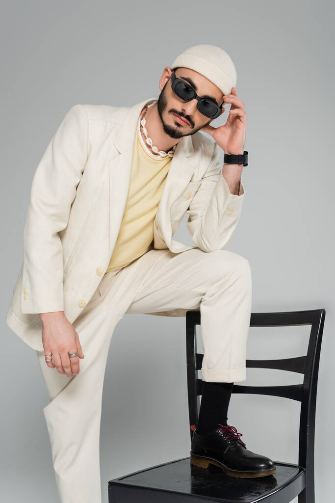 Μοντέρνος γκέι άντρας με γυαλιά ηλίου και κοστούμι που ποζάρει κοντά σε καρέκλα απομονωμένη σε γκρι   - Φωτογραφία, εικόνα