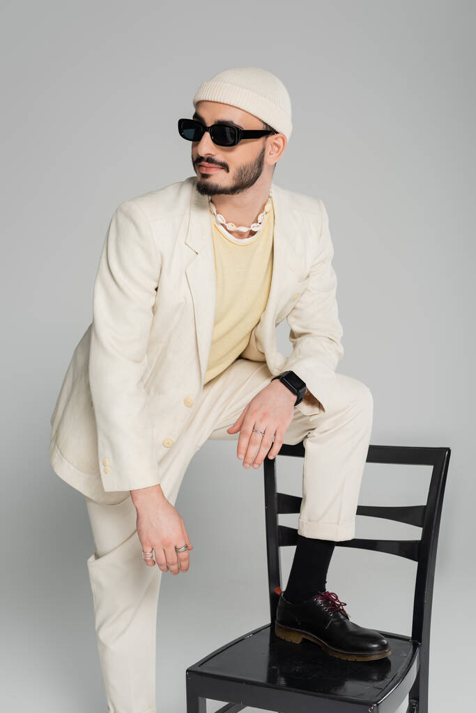 alla moda e barbuto gay uomo in abito in piedi vicino sedia isolato su grigio   - Foto, immagini