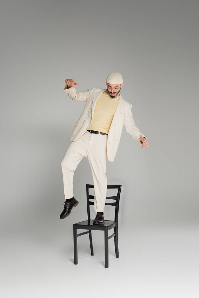 Стильний гей-чоловік у бежевому костюмі та капелюсі, що стоїть на стільці на сірому фоні
  - Фото, зображення