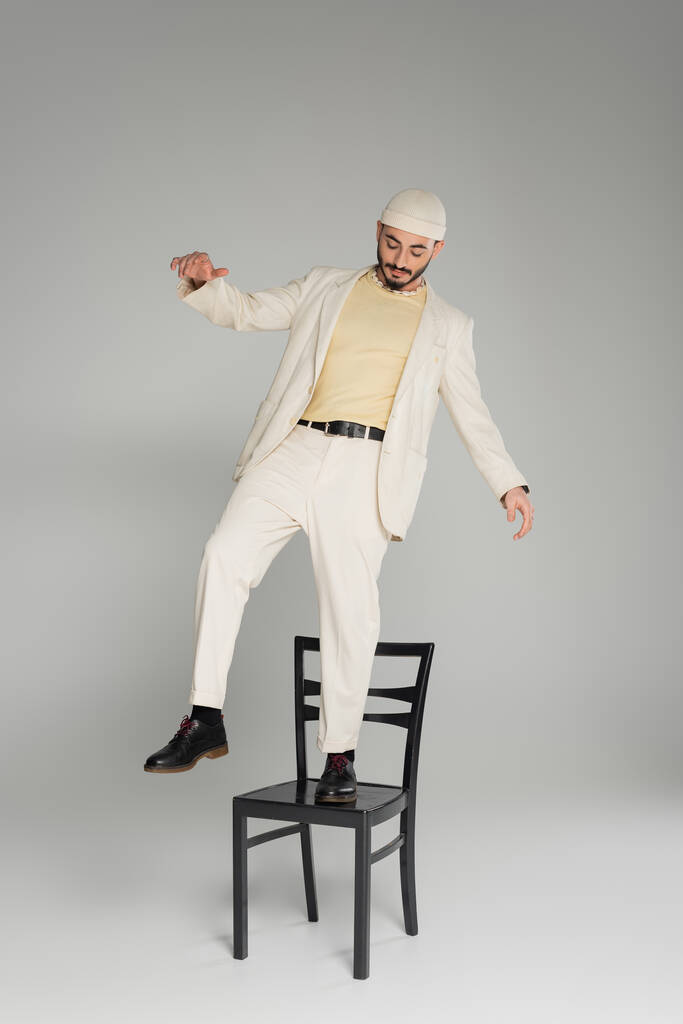 Modischer homosexueller Mann in Anzug und beigem Hut steht auf einem Stuhl vor grauem Hintergrund  - Foto, Bild