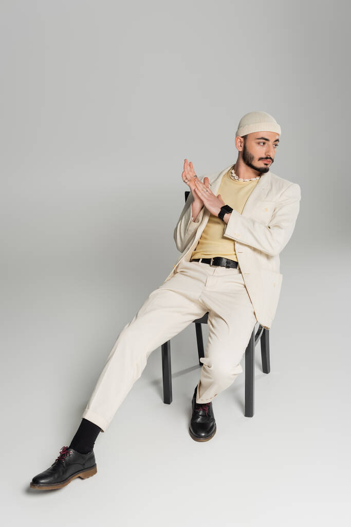 Trendy gay man in beige hat and suit sitting on chair on grey background  - Photo, Image