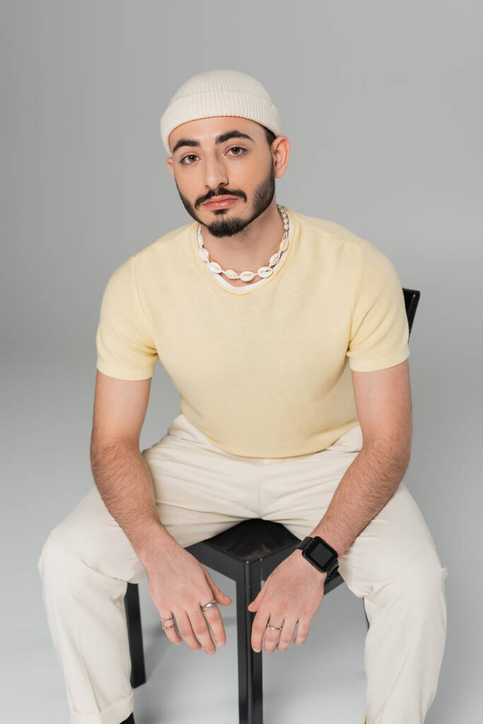Muodikas homo mies hatussa katselee kameraa istuessaan tuolilla harmaalla pohjalla  - Valokuva, kuva