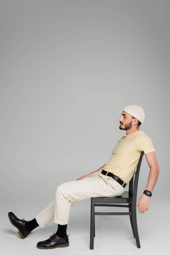 Seitenansicht eines bärtigen homosexuellen Mannes mit beigem Hut, der auf einem Stuhl auf grauem Hintergrund sitzt  - Foto, Bild
