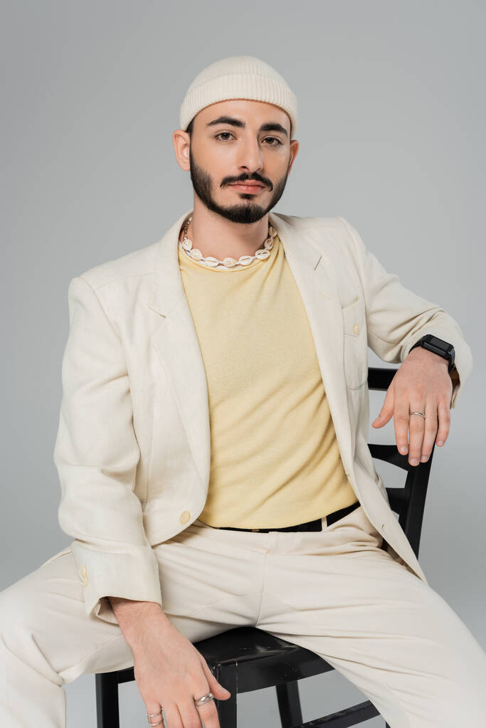 barbu homosexuel homme en costume ad coquillage collier assis sur chaise isolé sur gris   - Photo, image
