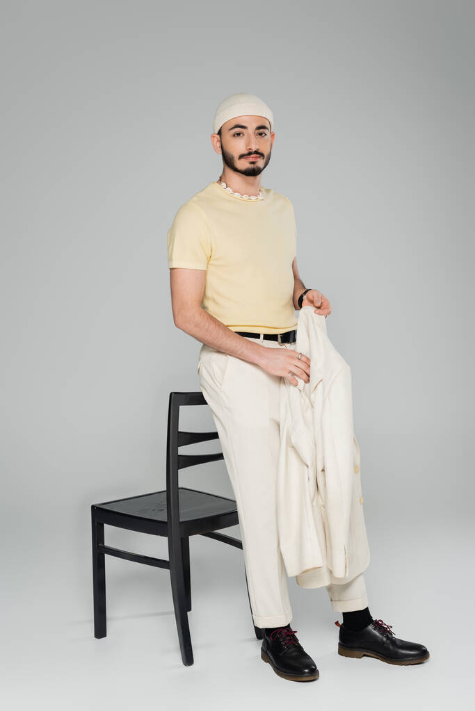 Volle Länge des trendigen homosexuellen Mannes mit Hut und Jacke in der Nähe des Stuhls auf grauem Hintergrund  - Foto, Bild
