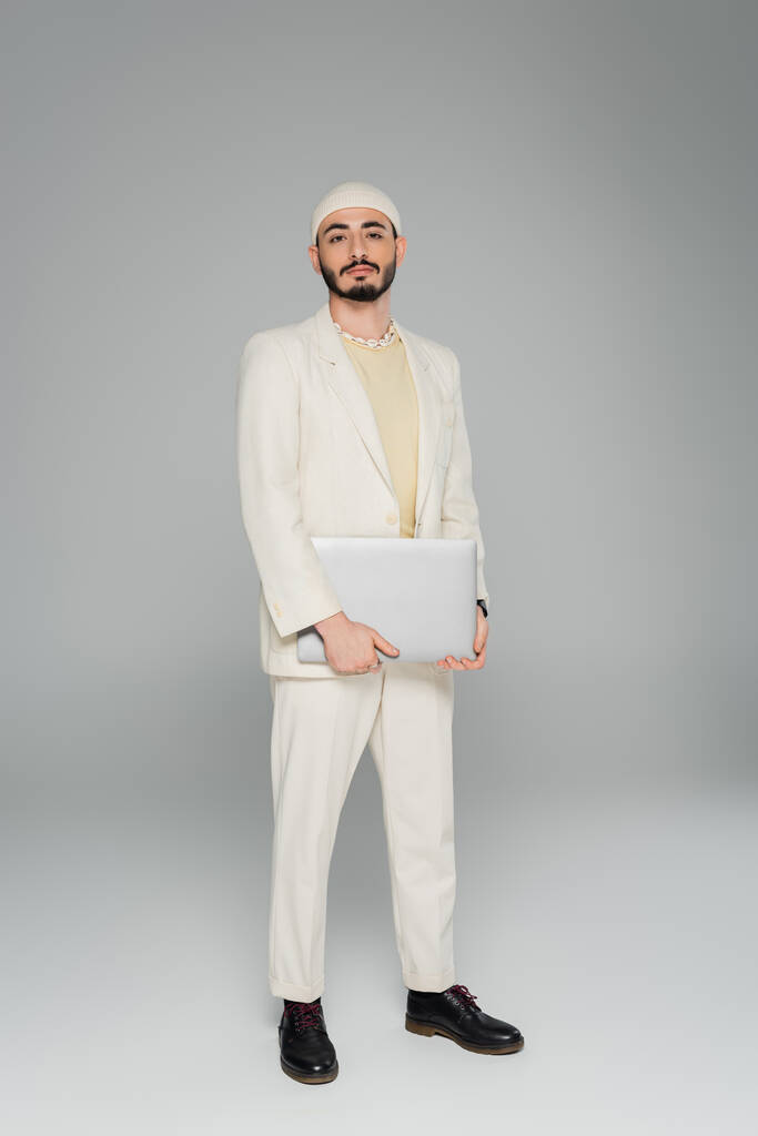 Stijlvolle gay man in beige pak holding laptop op grijze achtergrond  - Foto, afbeelding