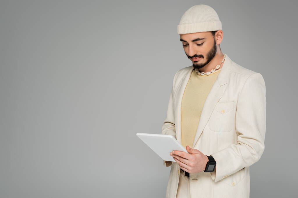 Young gay man in beige suit and hat using digital tablet isolated on grey   - Photo, Image