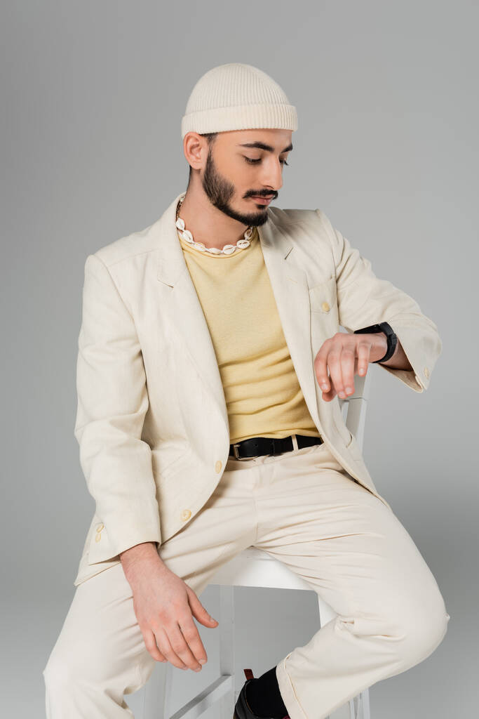 Stilvoller schwuler Mann im Anzug schaut auf intelligente Uhr, während er auf einem Stuhl sitzt, isoliert auf grau   - Foto, Bild