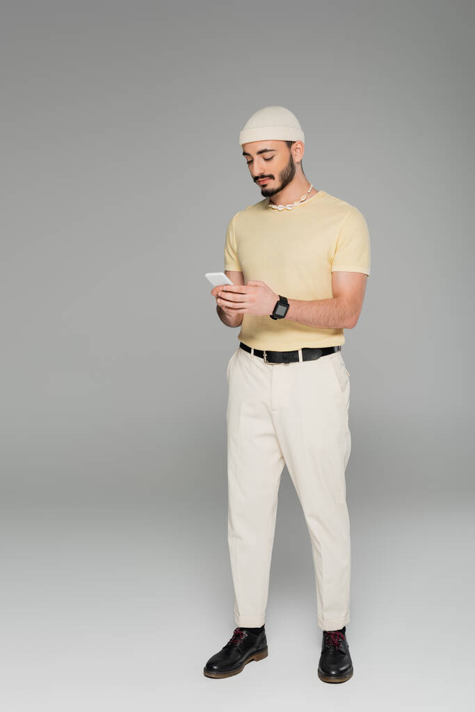 na moda gay homem no chapéu usando celular enquanto em pé no cinza fundo  - Foto, Imagem