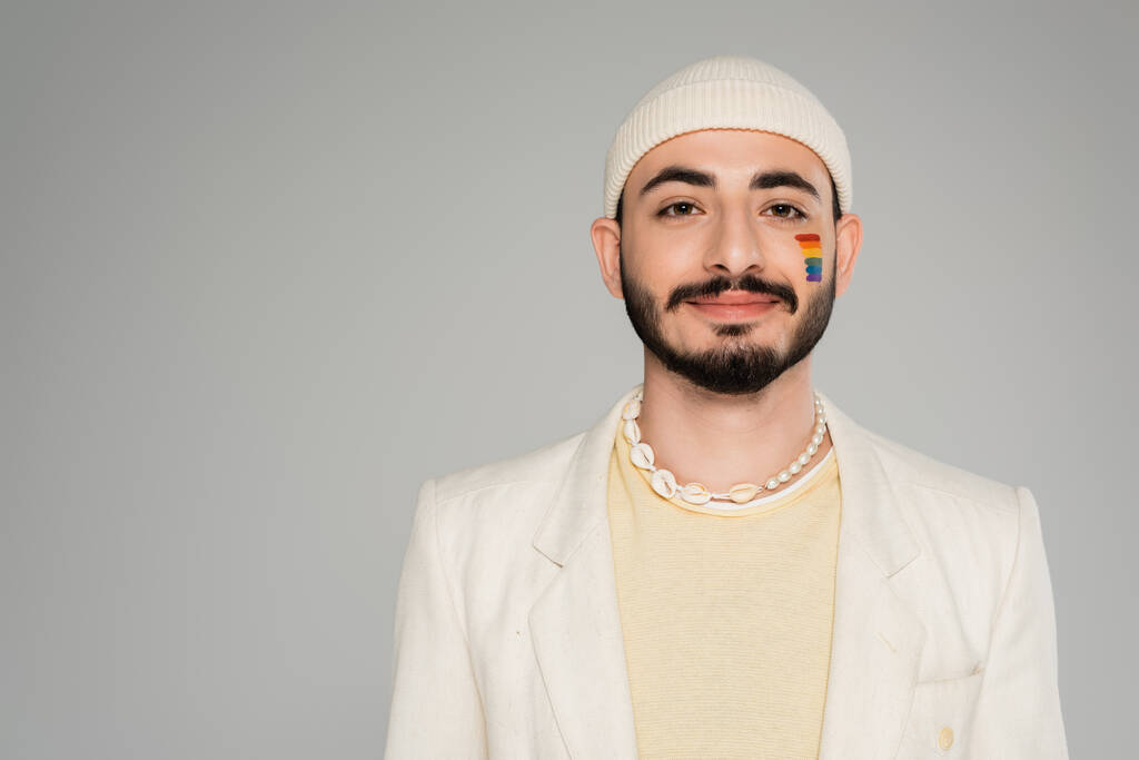 Portrét stylového gaye s vlajkou lgbt na tváři při pohledu na kameru izolovanou na šedi   - Fotografie, Obrázek