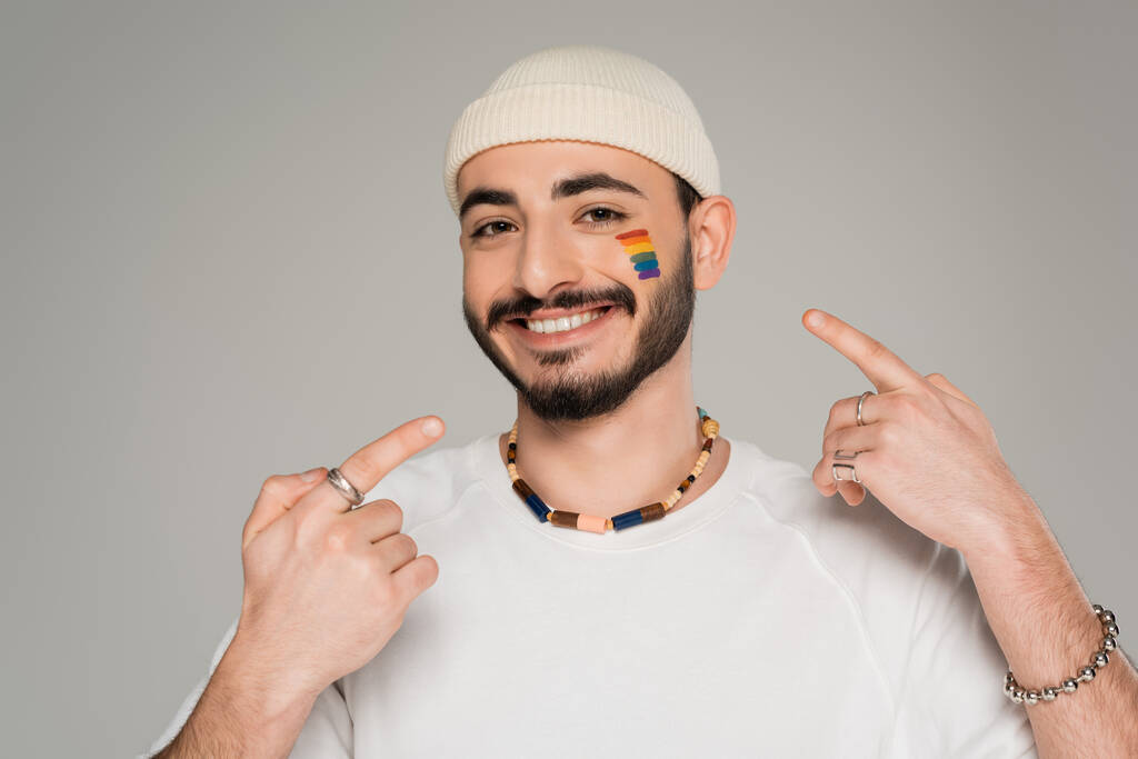 Улыбающийся гомосексуалист в шляпе, указывающий на флаг lgbt на щеке, изолированный от серого   - Фото, изображение