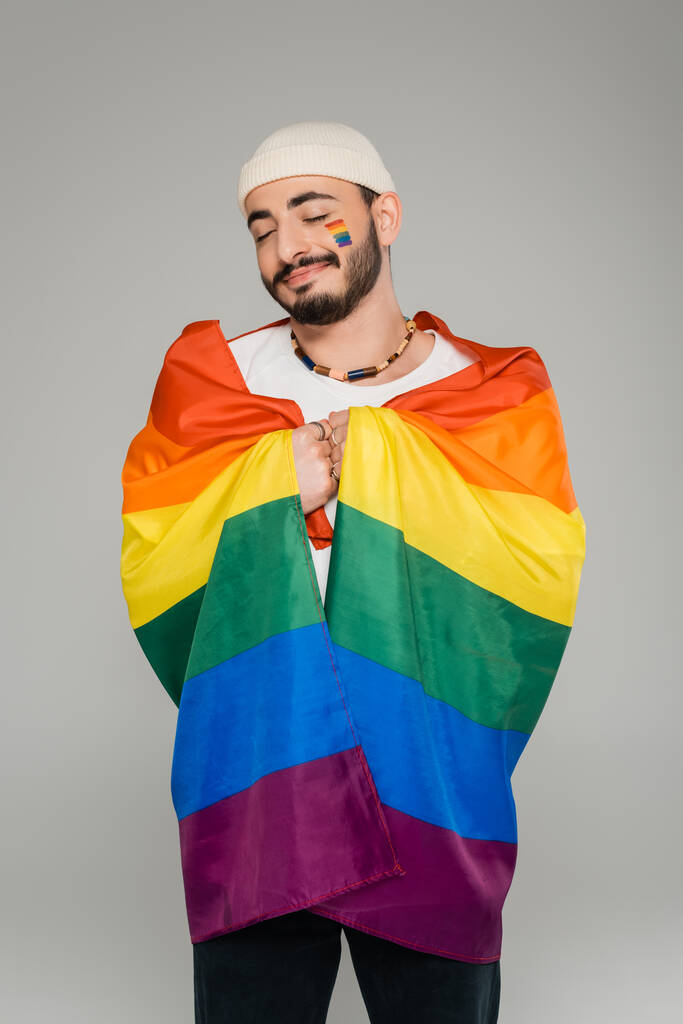 Joyful gay man holding lgbt flag and closing eyes isolated on grey   - Photo, Image