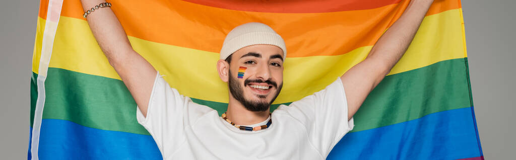 allegro gay uomo in cappello tenendo lgbt bandiera e guardando fotocamera isolato su grigio banner  - Foto, immagini